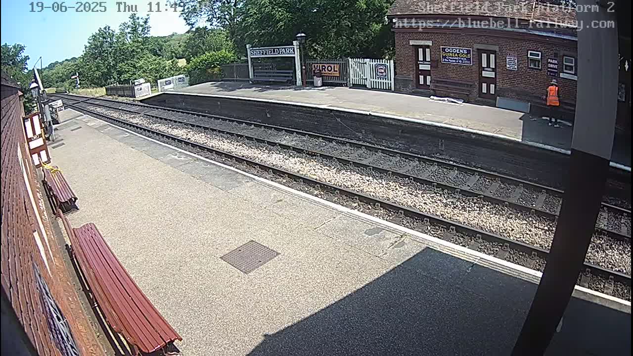 A sunny view of a train station platform with two benches, railway tracks, and a person in an orange vest standing near a brick building.