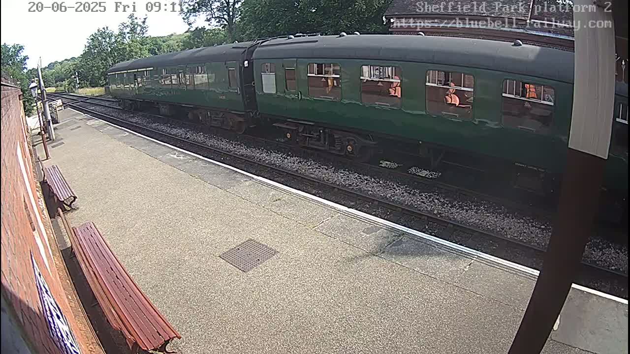 A long, dark-green passenger train sits at a station platform on a sunny day.