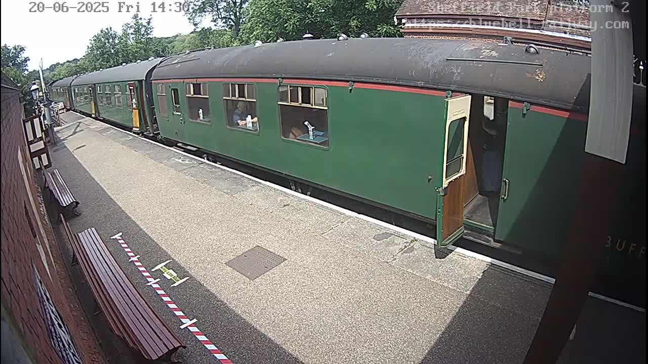 A long, dark-green passenger train is stopped at a station platform on a sunny day.