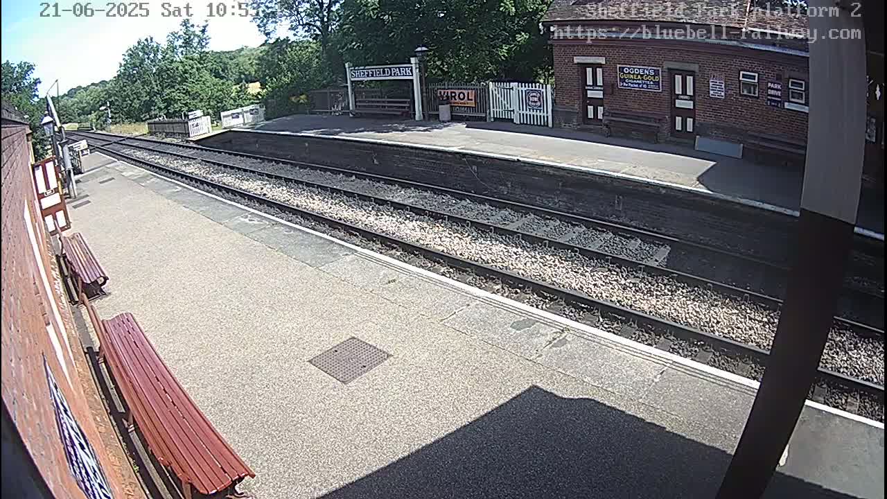 A sunny view of a train station platform with two parallel sets of train tracks, benches, and a brick building.