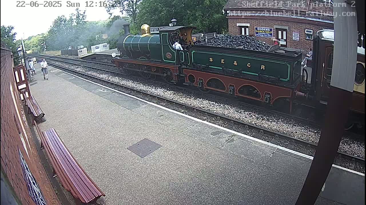 A steam train carrying coal is at a station platform on a partly cloudy day.