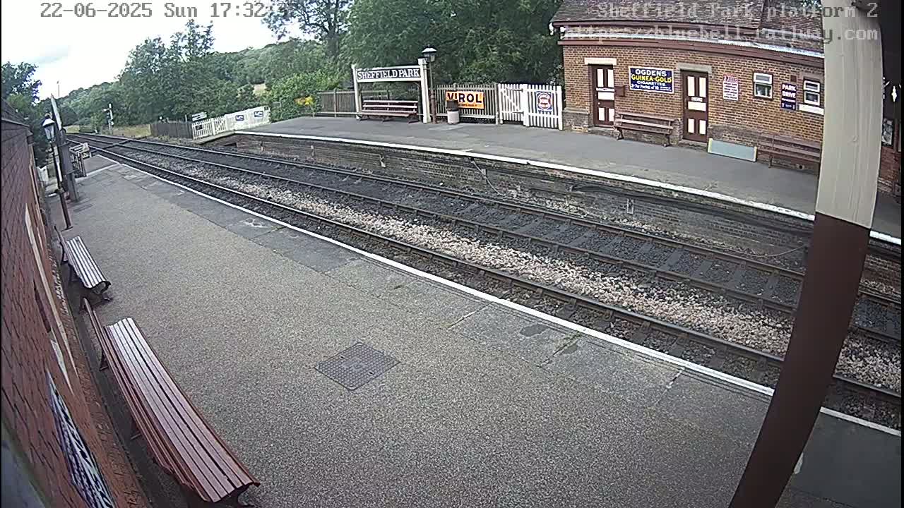 A train platform with two benches and railway tracks stretches into the distance under a partly cloudy sky.
