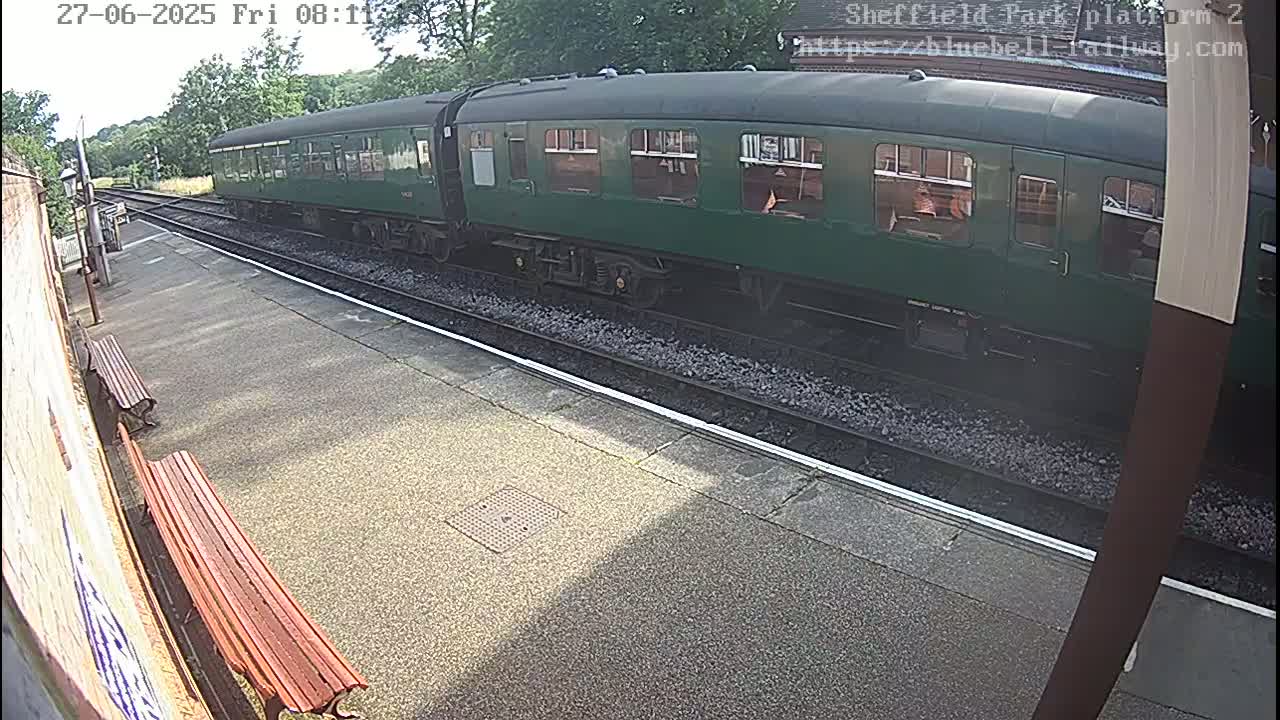 A long, dark-green passenger train sits at a station platform on a sunny day.
