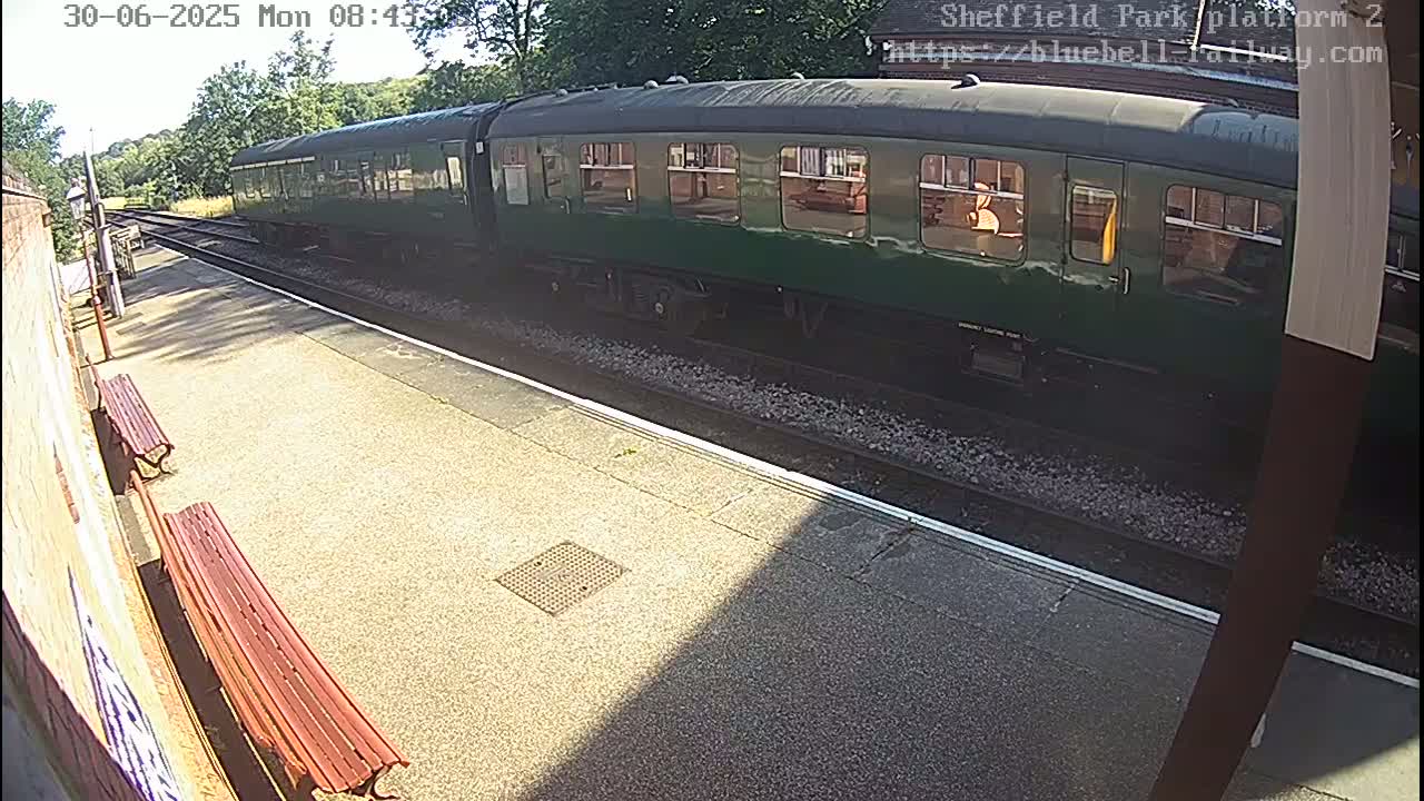 A dark-green passenger train is at a sunny train station platform.