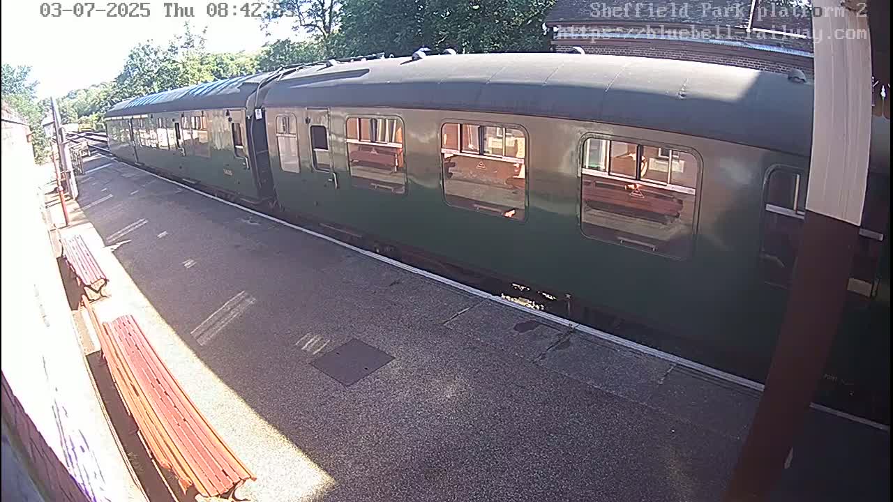 A long, dark green passenger train sits at a station platform on a sunny day.