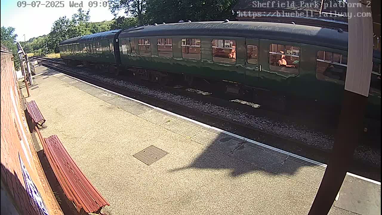 A long, dark green passenger train sits at a station platform on a sunny day.