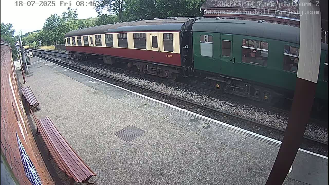 A red and cream passenger train car coupled to a green passenger car sits at a train station on a partly cloudy day.