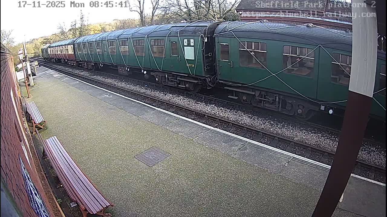 A long train with green and maroon carriages is stopped at a station platform, featuring benches and a brick wall, under an overcast sky.