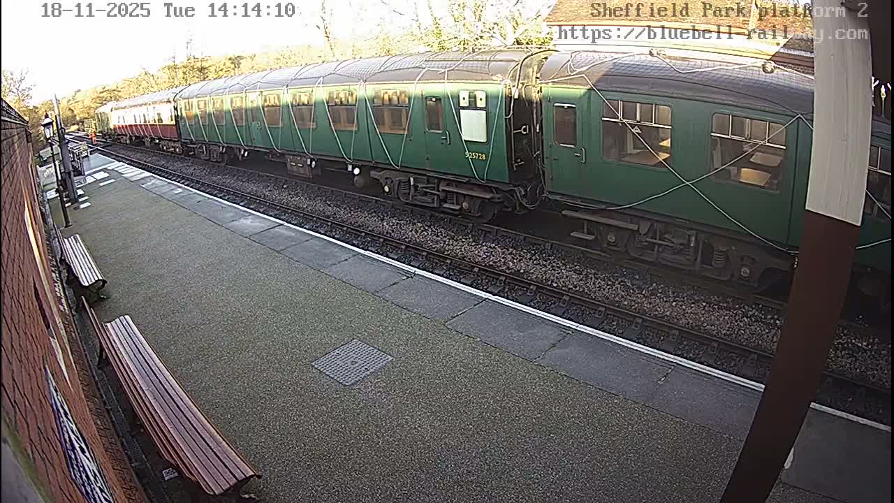 A long train with green and maroon carriages is stopped at a station platform, featuring benches and a brick wall, under an overcast sky.