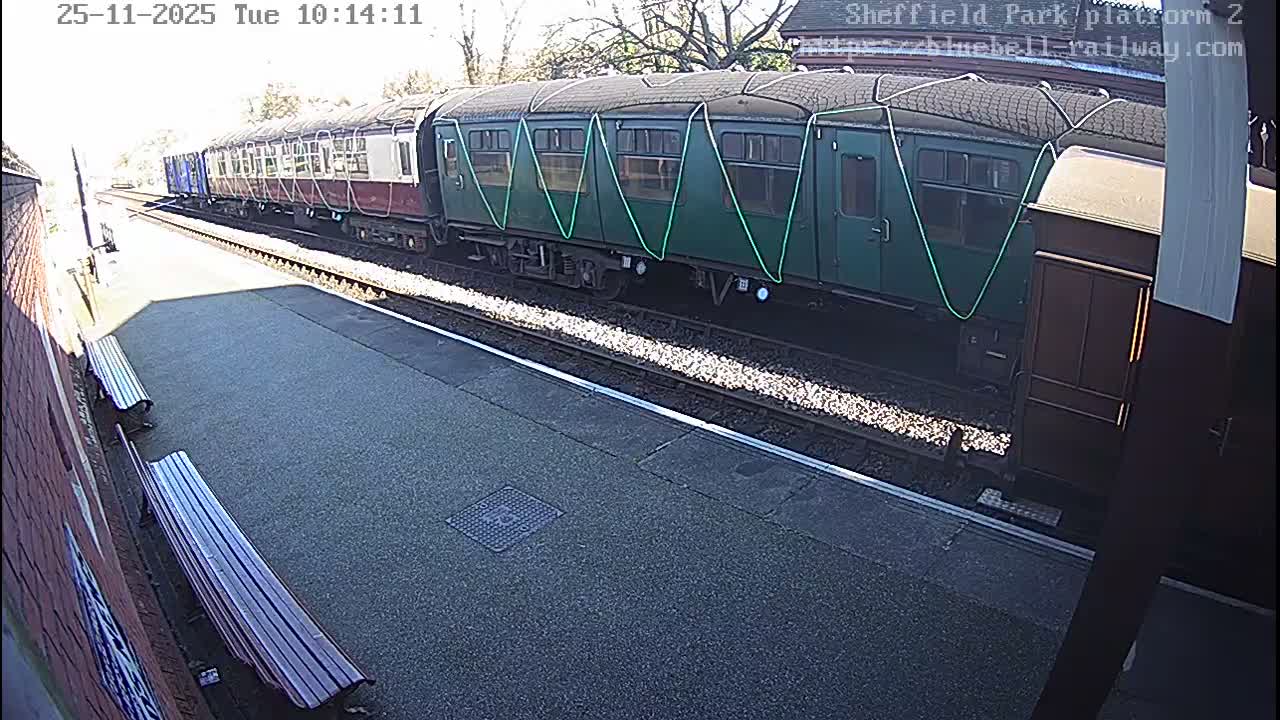 A multi-car train, featuring green and maroon carriages, is visible alongside a station platform equipped with benches under bright, sunny skies.