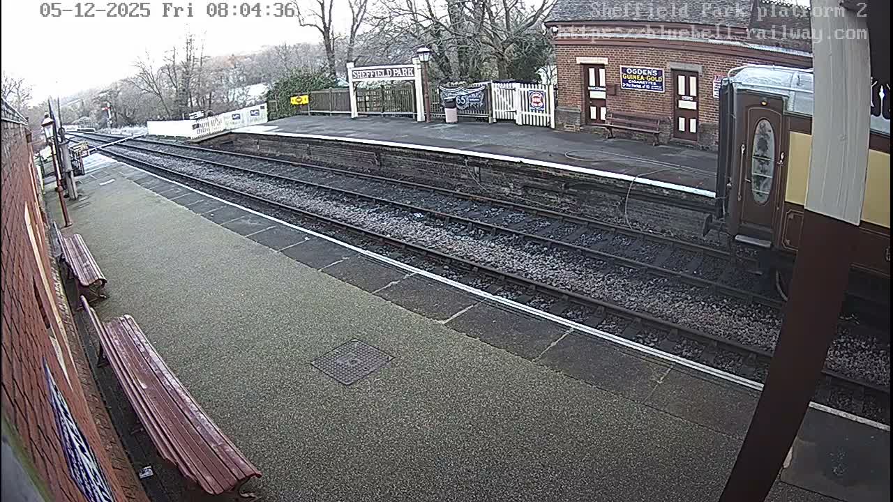 An empty Sheffield Park railway station is seen under an overcast sky, featuring tracks, platforms with benches, a brick station building, and a partially visible train carriage.