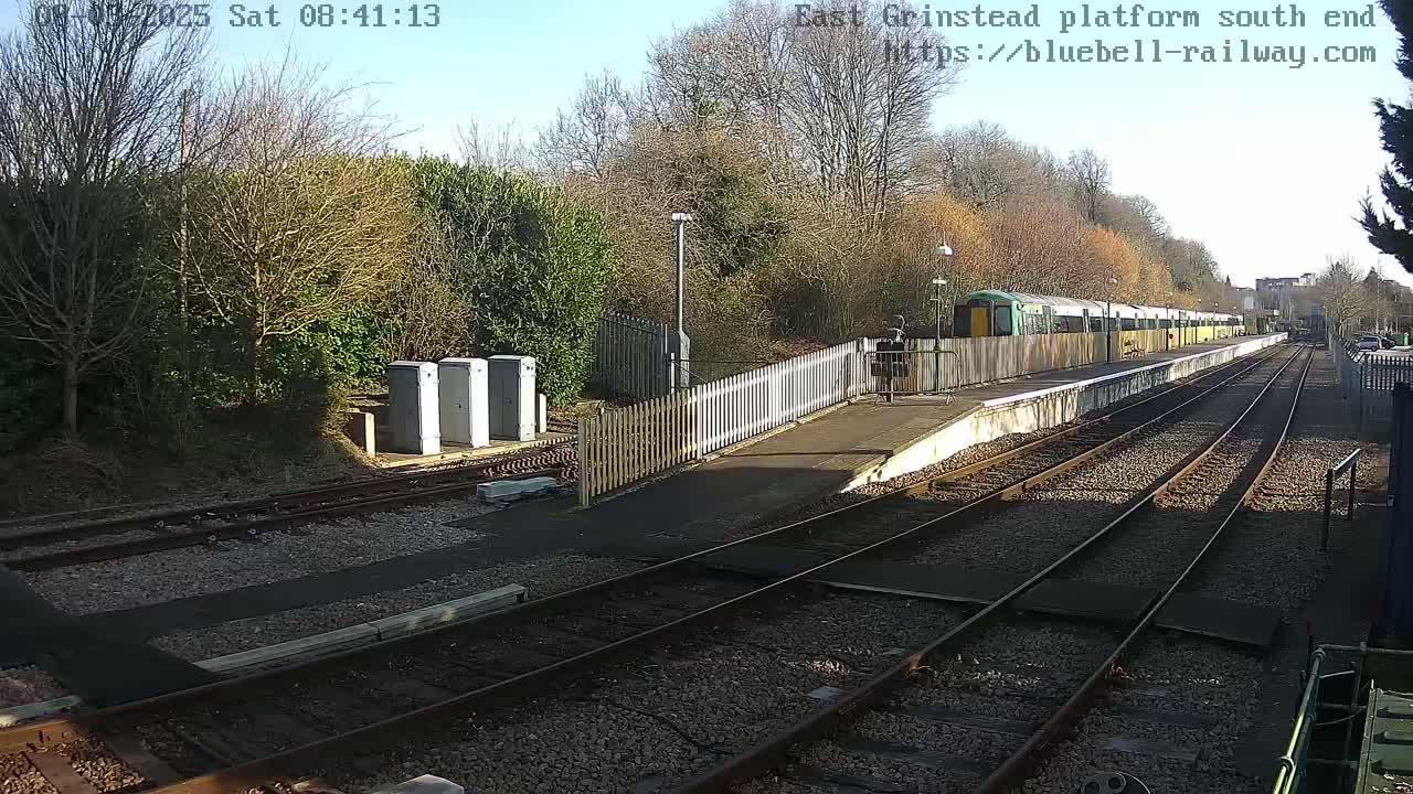 A sunny day at a train station shows a long train stopped at a platform with multiple tracks and a wooden fence, surrounded by shrubs and trees.