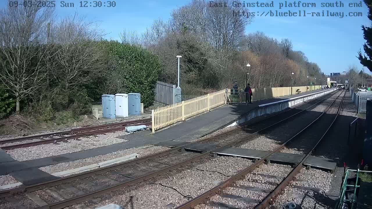 A sunny day at a train station platform with a few people, some small buildings, and a wooden fence bordering the tracks.