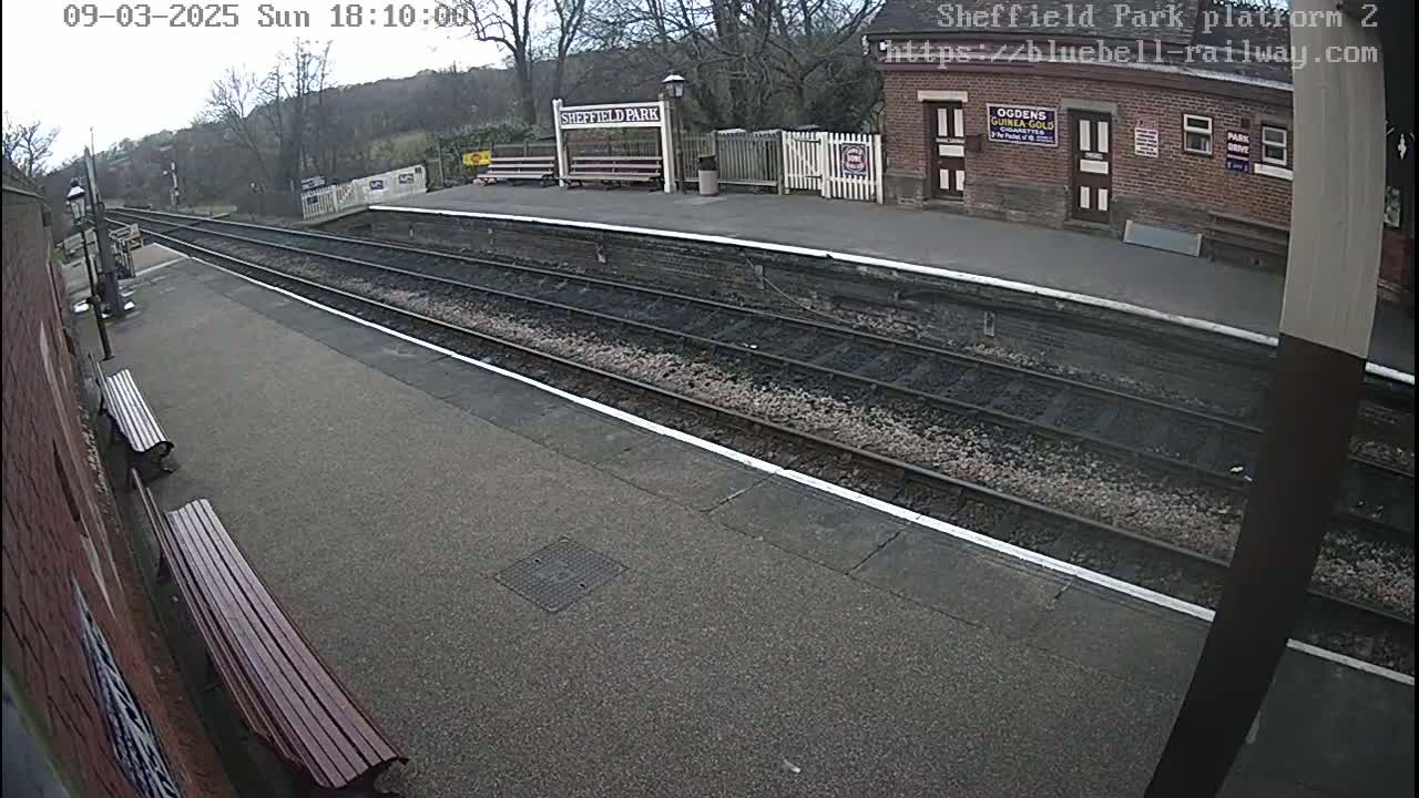 An overcast day view of a train platform with two benches, a brick building, and multiple train tracks.