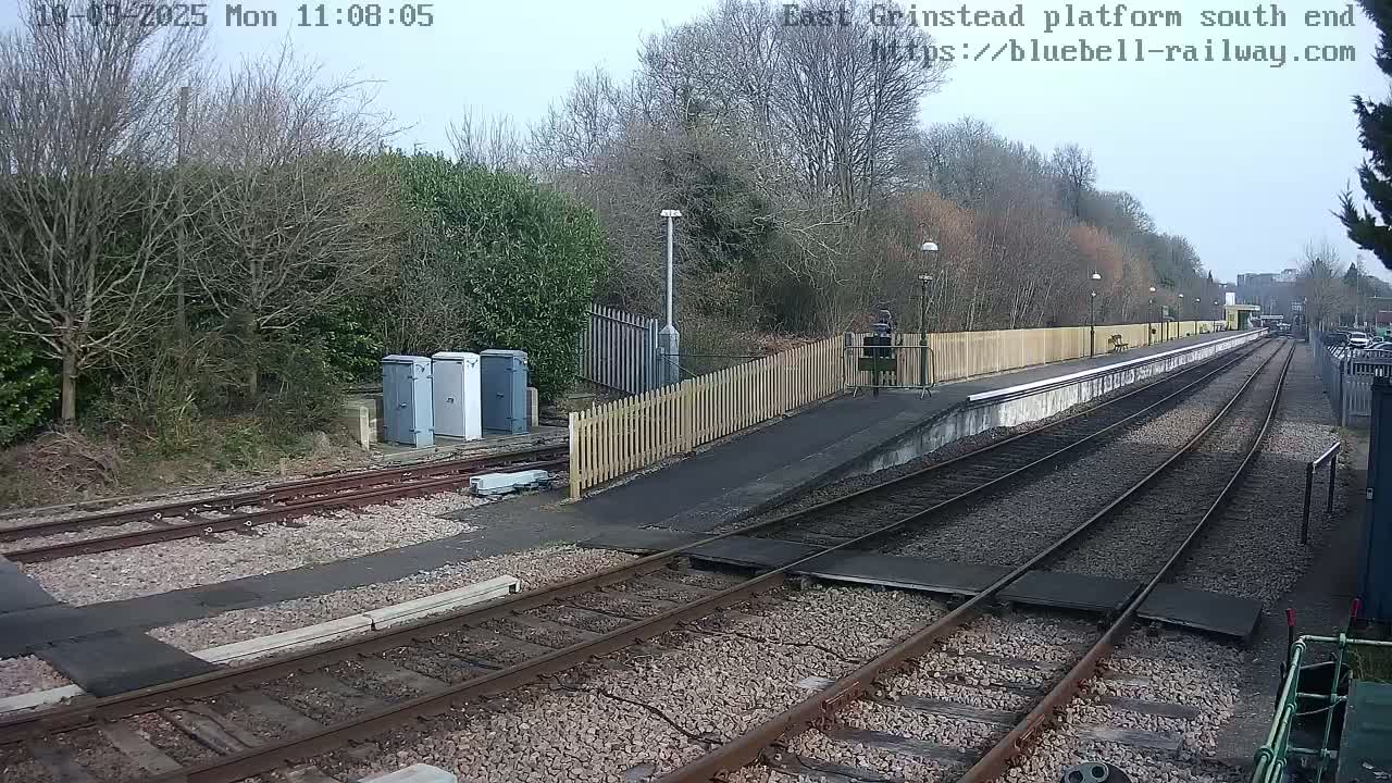 A view of a train platform on a mostly clear day, with bare trees and shrubs surrounding the railway tracks.