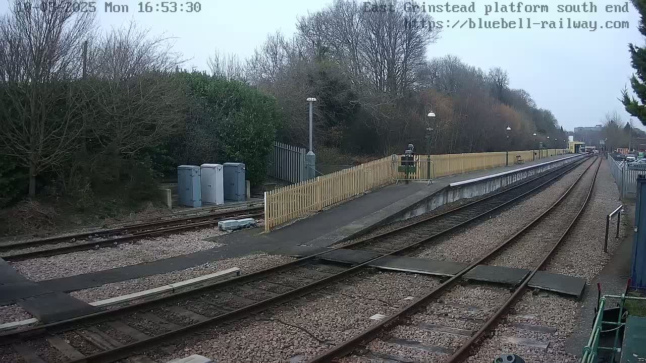 A view of a train platform on an overcast day, showing multiple train tracks extending into the distance, bordered by a wooden fence and bare trees.