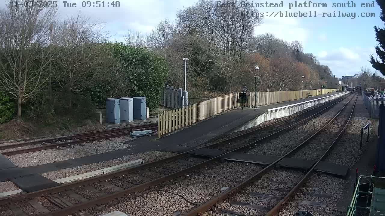 A mostly sunny view of a train platform with two sets of tracks leading away from a low, wooden fence, and a few bare trees and shrubs visible in the background.