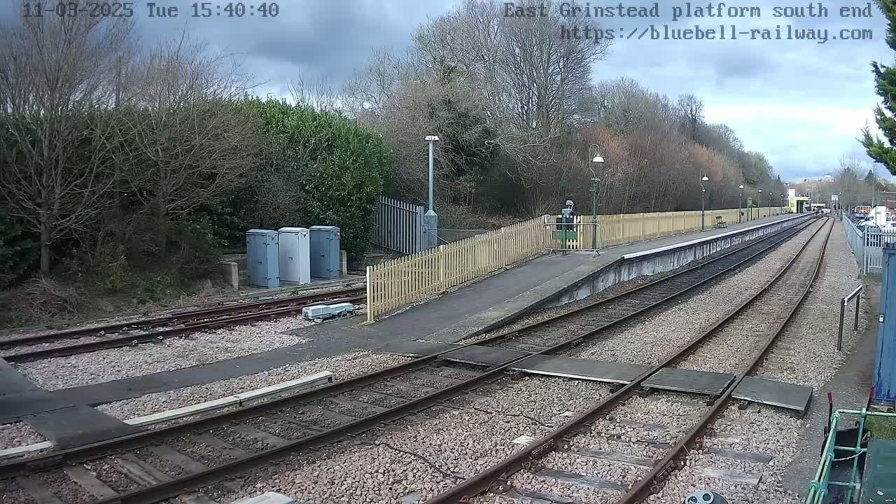 A train platform at the south end, with overcast skies, shows several train tracks extending into the distance, bordered by a wooden fence and trees.