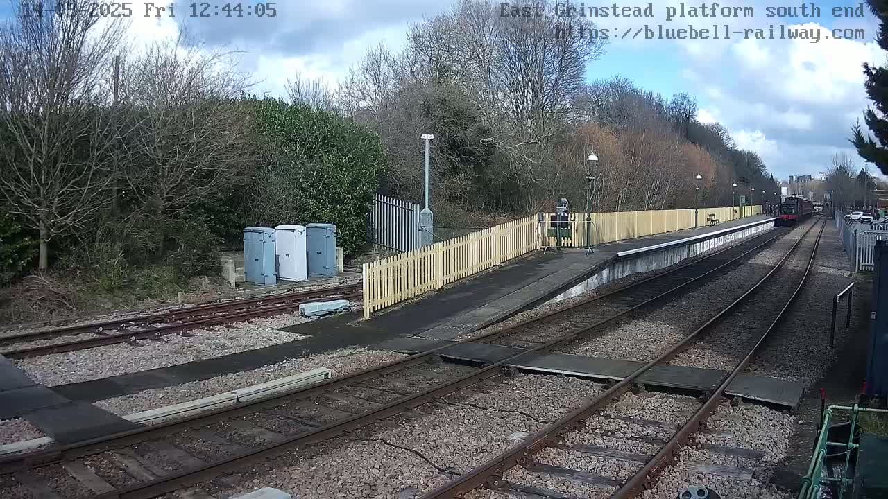 A train departs from a railway platform under a partly cloudy sky, passing a wooden fence and several utility boxes.