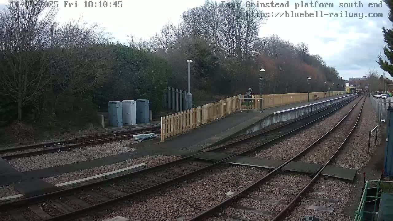A view of a train platform on an overcast day, showing train tracks extending into the distance, alongside a wooden fence and a few buildings.