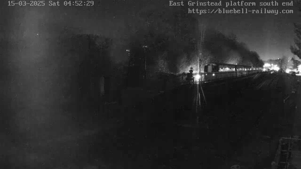 A nighttime, low-light view shows a train departing from a platform, emitting smoke.