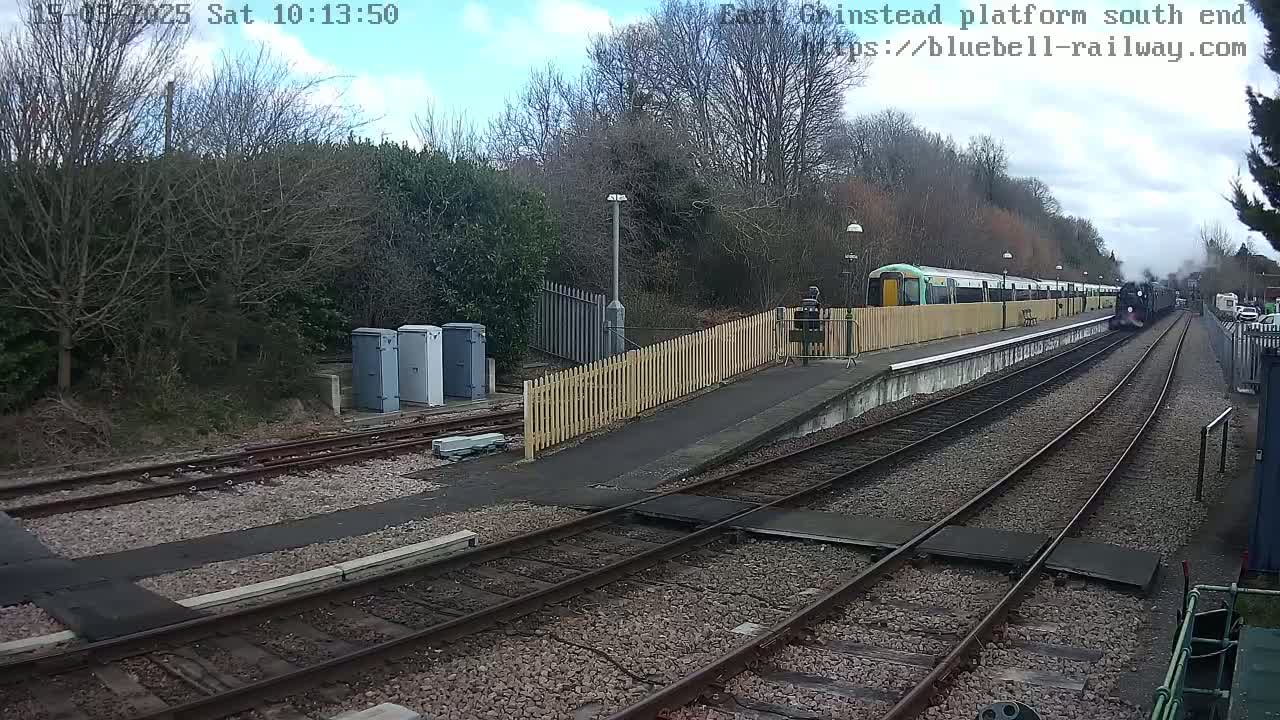 A steam train is departing a railway platform on a partly cloudy day, passing a modern electric multiple unit train.