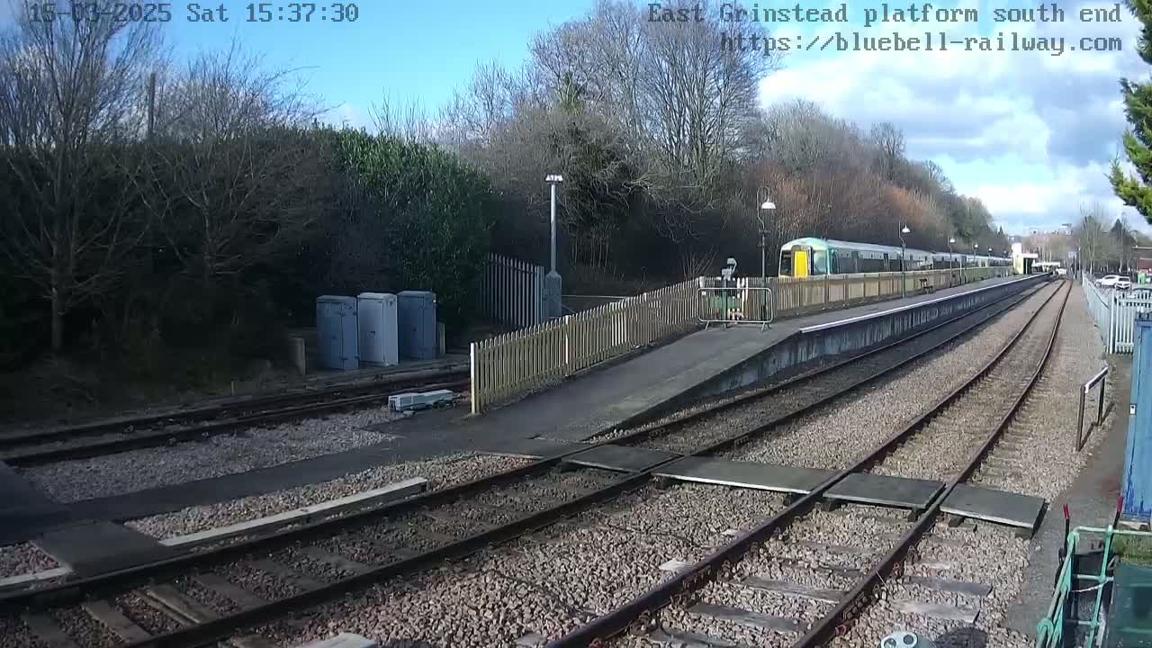 A train is stopped at a railway platform on a sunny day, with trees and shrubs visible in the background.
