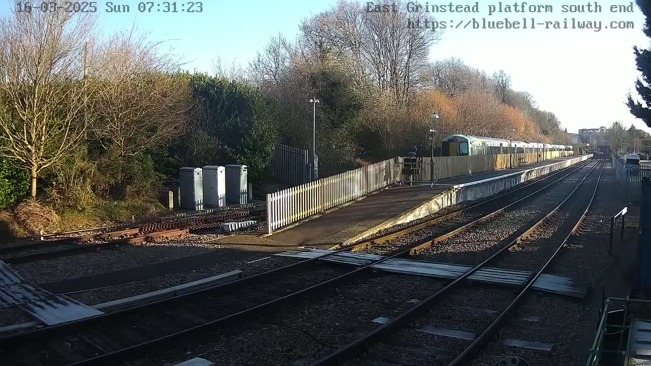 A sunny day at a train station shows a train at the platform, multiple sets of tracks, and a white picket fence bordering the platform.
