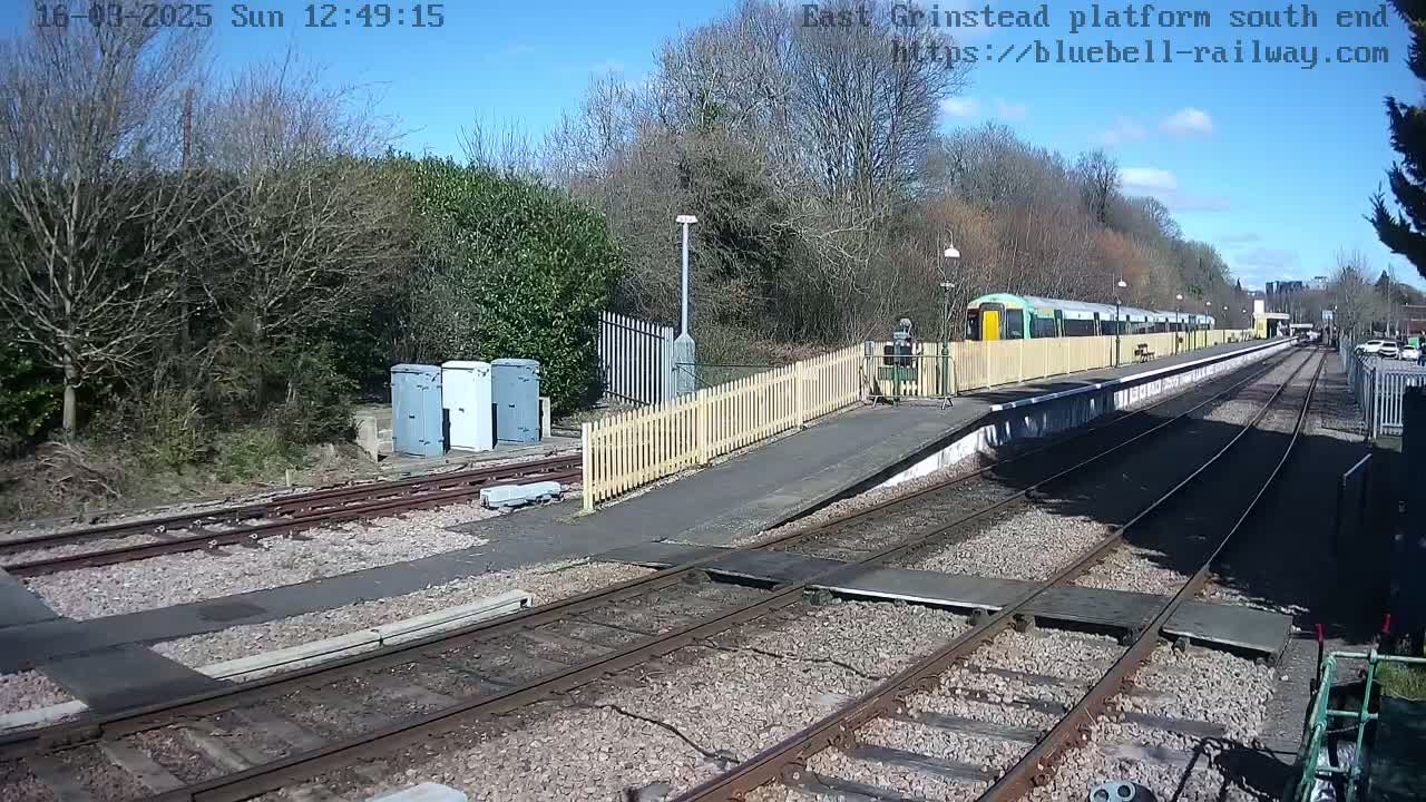 A train sits at a station platform on a sunny day, with bare trees and shrubs surrounding the tracks.