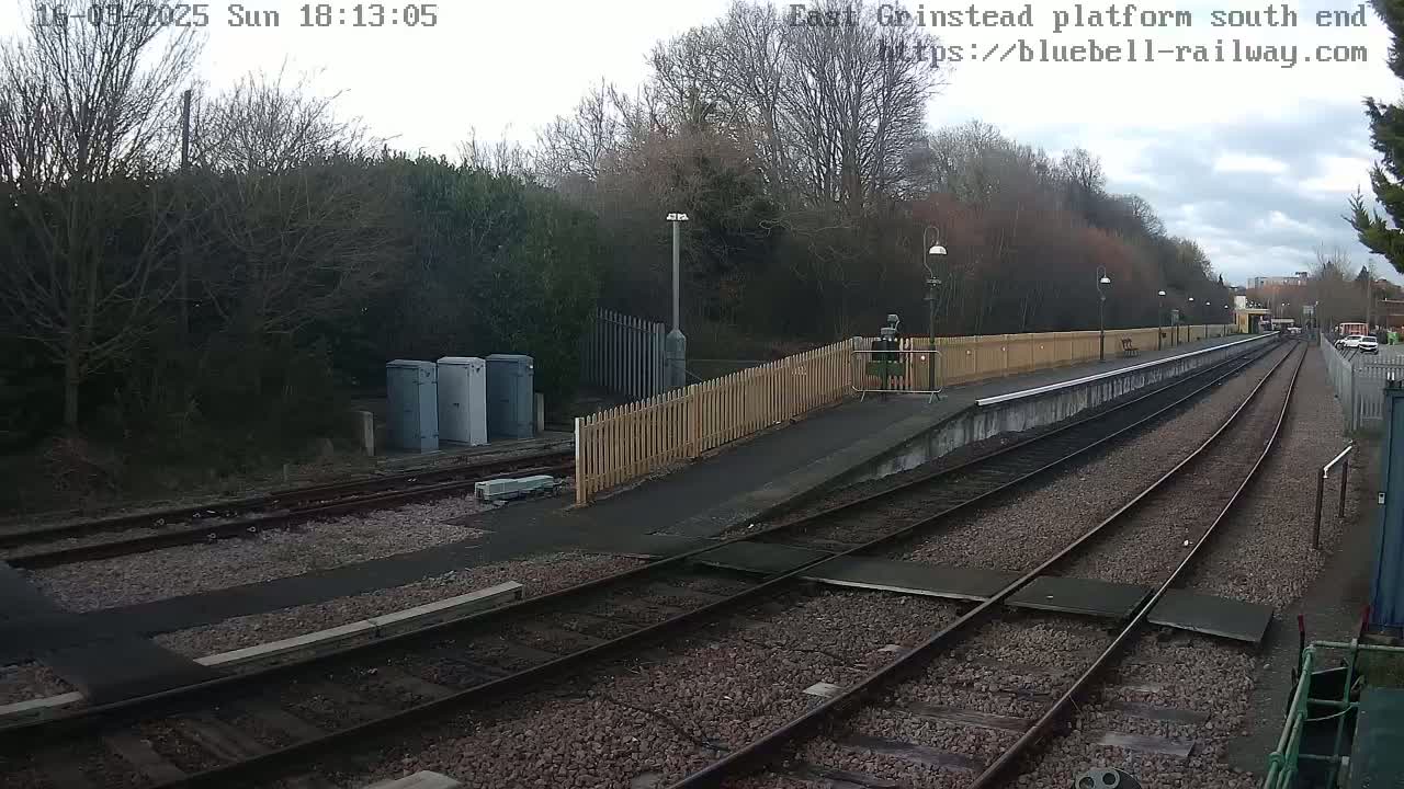 An overcast day at a train station with two sets of train tracks extending into the distance, bordered by a wooden fence and sparsely populated with trees.