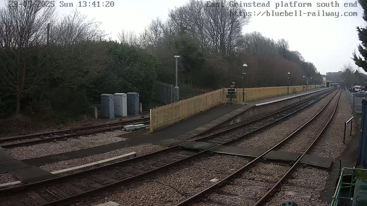 A train platform with two sets of tracks extends into the distance under an overcast sky, bordered by a wooden fence and a line of trees.