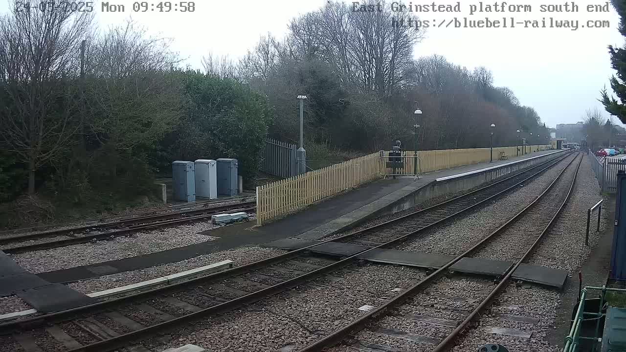 A view of a train platform on an overcast day, showing railway tracks extending into the distance, bordered by a wooden fence and trees.