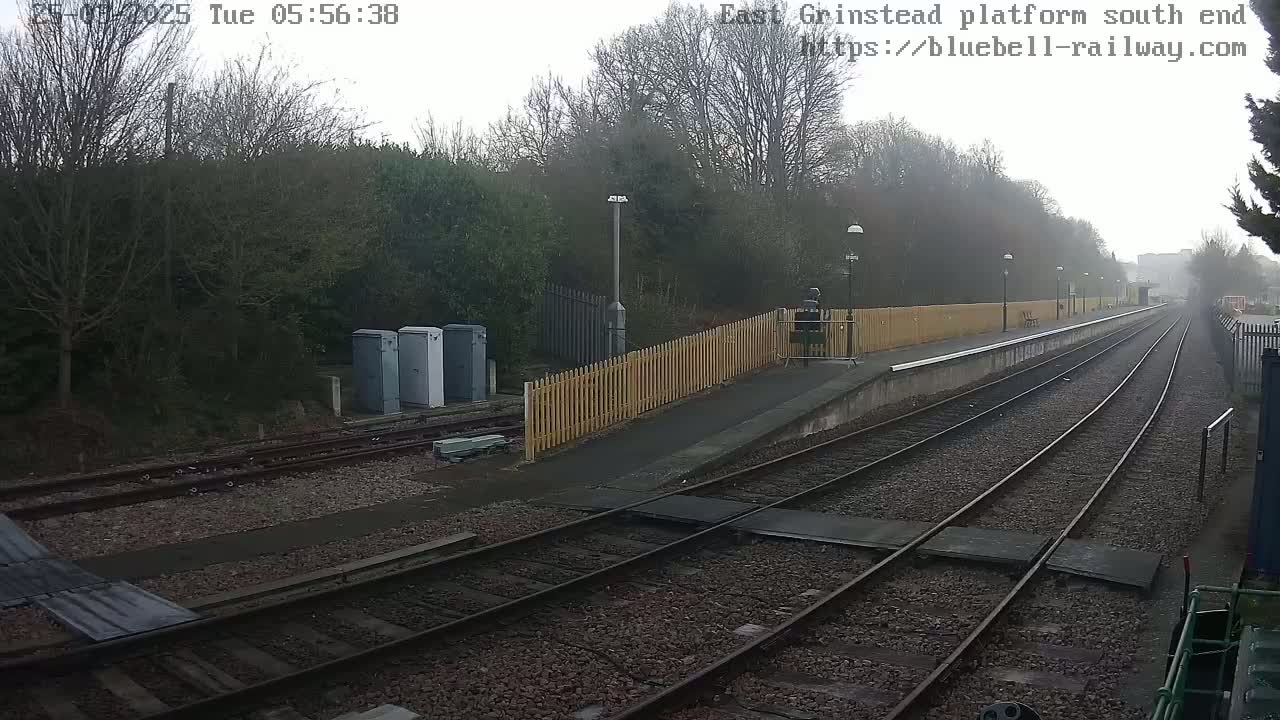 A foggy view of a train platform with two sets of tracks extending into the distance, bordered by a wooden fence and surrounded by trees.