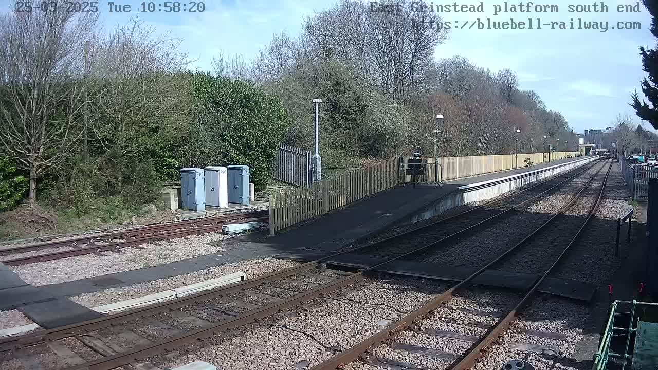 A train platform at a railway station on a sunny day, with multiple train tracks extending into the distance, surrounded by trees and shrubs.