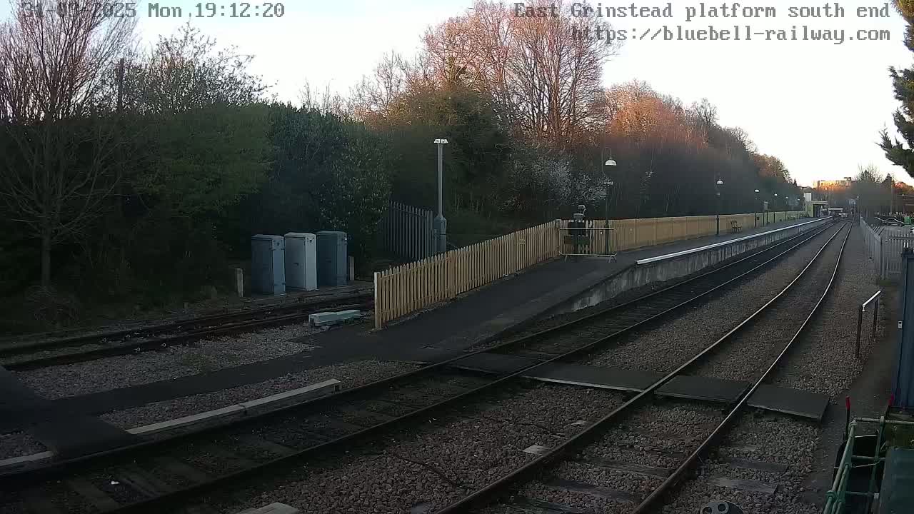 A train platform at dusk, with two sets of train tracks extending into the distance, bordered by a wooden fence and trees.
