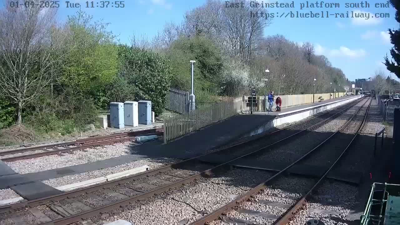 A sunny day at a train station with a few people waiting on the platform, alongside train tracks.
