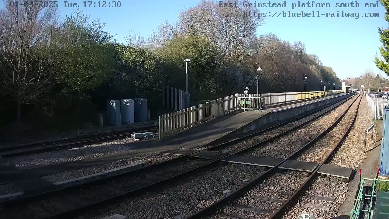 A sunny day at a train station platform with multiple train tracks, a wooden fence, and a small building.