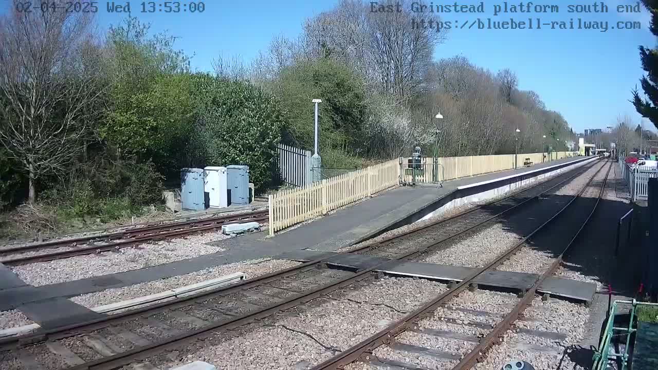 A sunny day at a train platform, showing two sets of train tracks leading to a small station bordered by a wooden fence and trees.