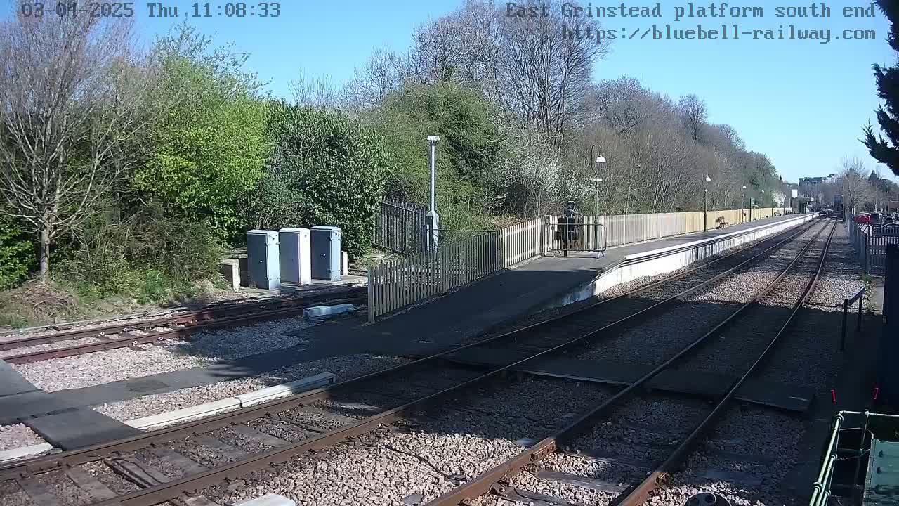 A sunny day at a train station platform, showing train tracks extending into the distance, alongside a low wooden fence and greenery.
