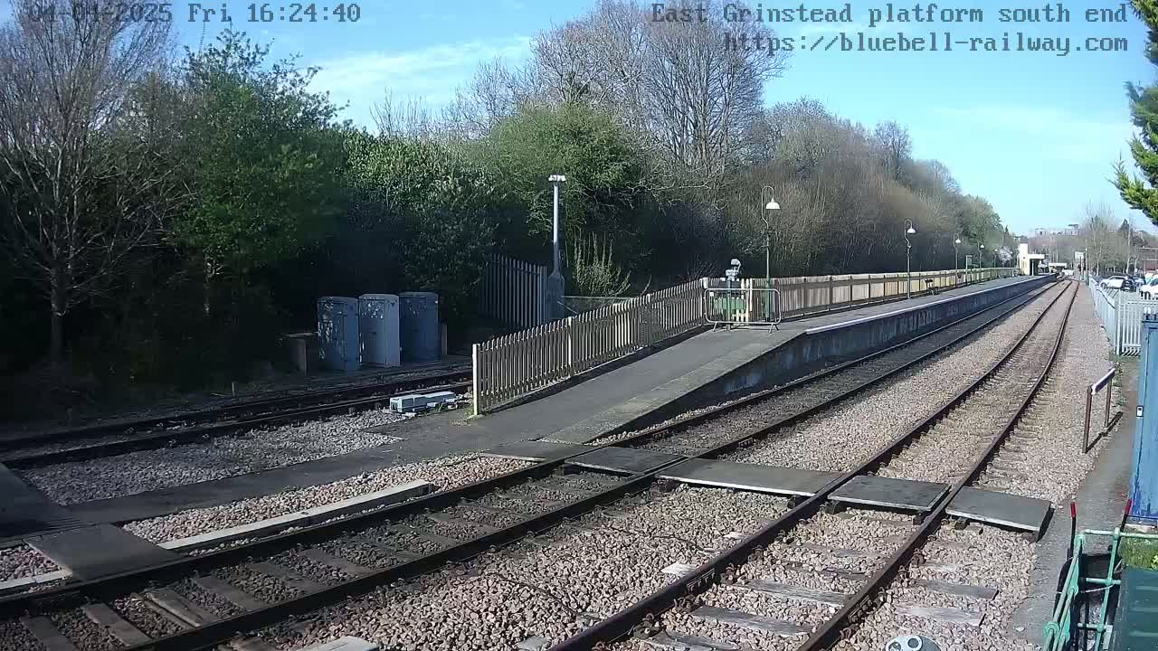 A sunny day at a train platform shows a view down two parallel sets of train tracks, bordered by a wooden fence, with some trees and shrubs in the background.
