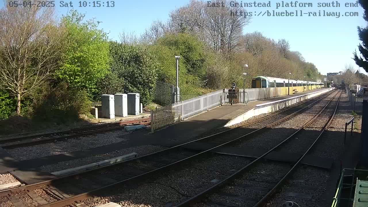 A sunny day at a train station shows a passenger train stopped at a platform, surrounded by trees and railway tracks.