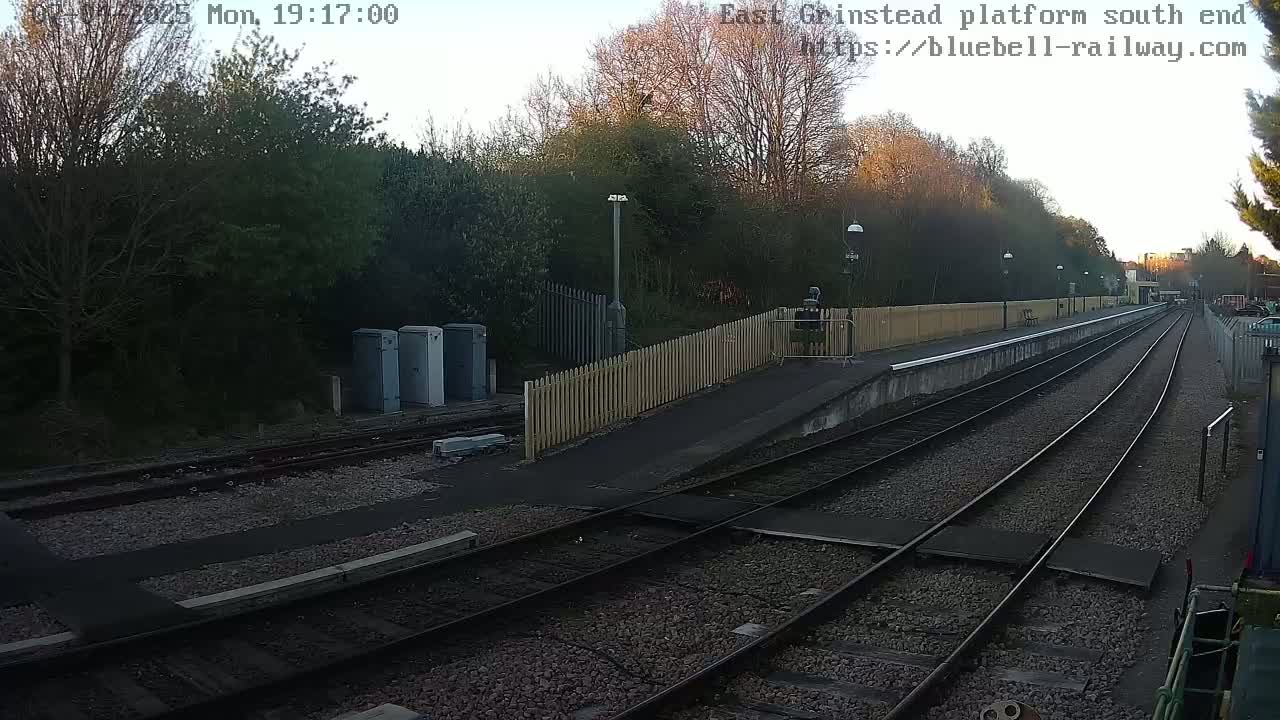 A train platform at dusk, with two sets of train tracks extending into the distance, bordered by a wooden fence and trees.