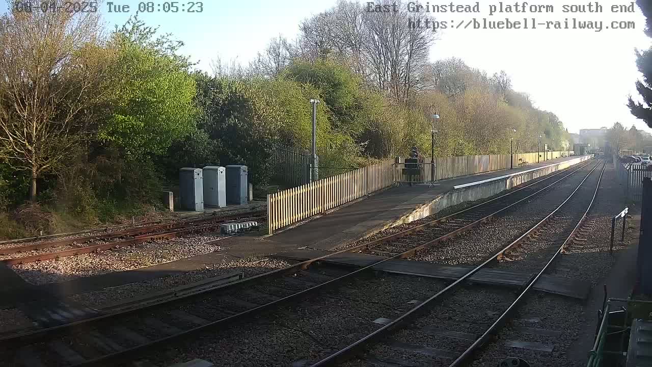 A sunny view of a train platform with two sets of tracks extending into the distance, bordered by a wooden fence and trees.