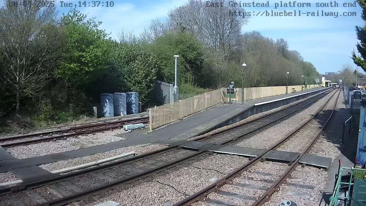 A view of a train platform on a sunny day, showing train tracks extending into the distance, bordered by a wooden fence and surrounded by trees.