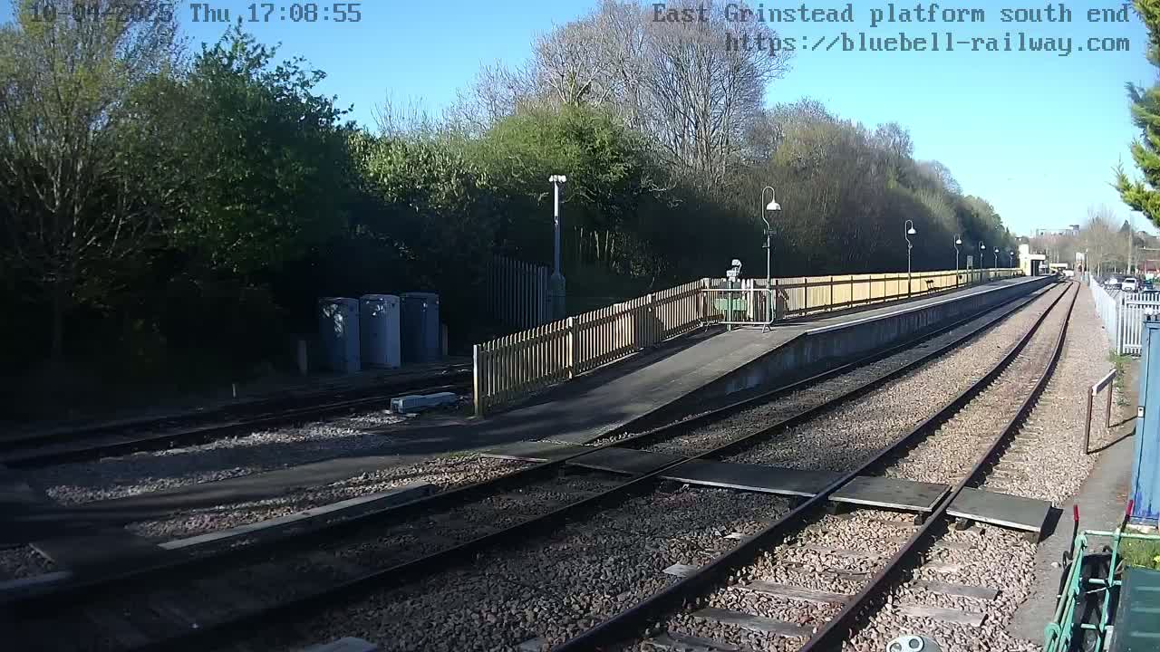A sunny view of a train platform with two sets of tracks, a wooden fence, and several light posts.