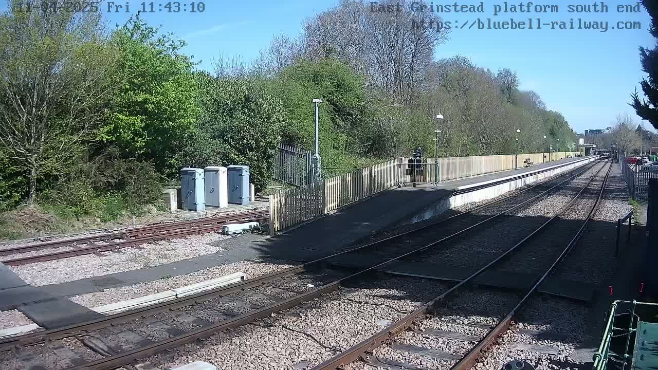 A sunny day at a train station platform shows train tracks extending into the distance, alongside a short platform bordered by a wooden fence and lush greenery.