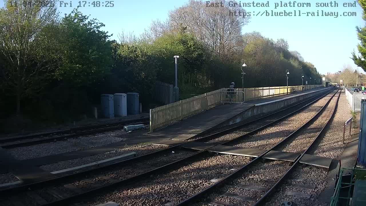 A view of a train platform on a sunny day, showing train tracks, a wooden fence, and some electrical boxes.
