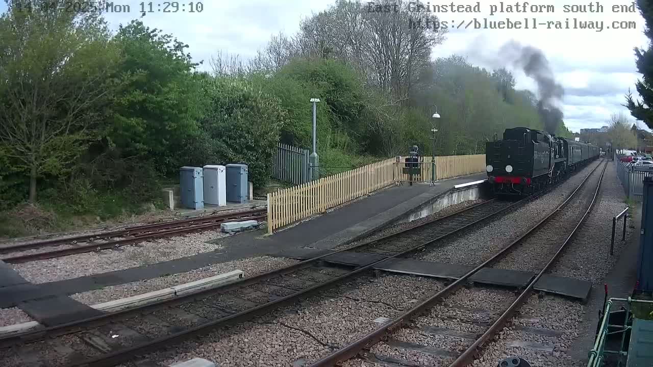 A dark-colored steam train is departing a railway station on an overcast day, passing alongside a wooden fence and several utility boxes.