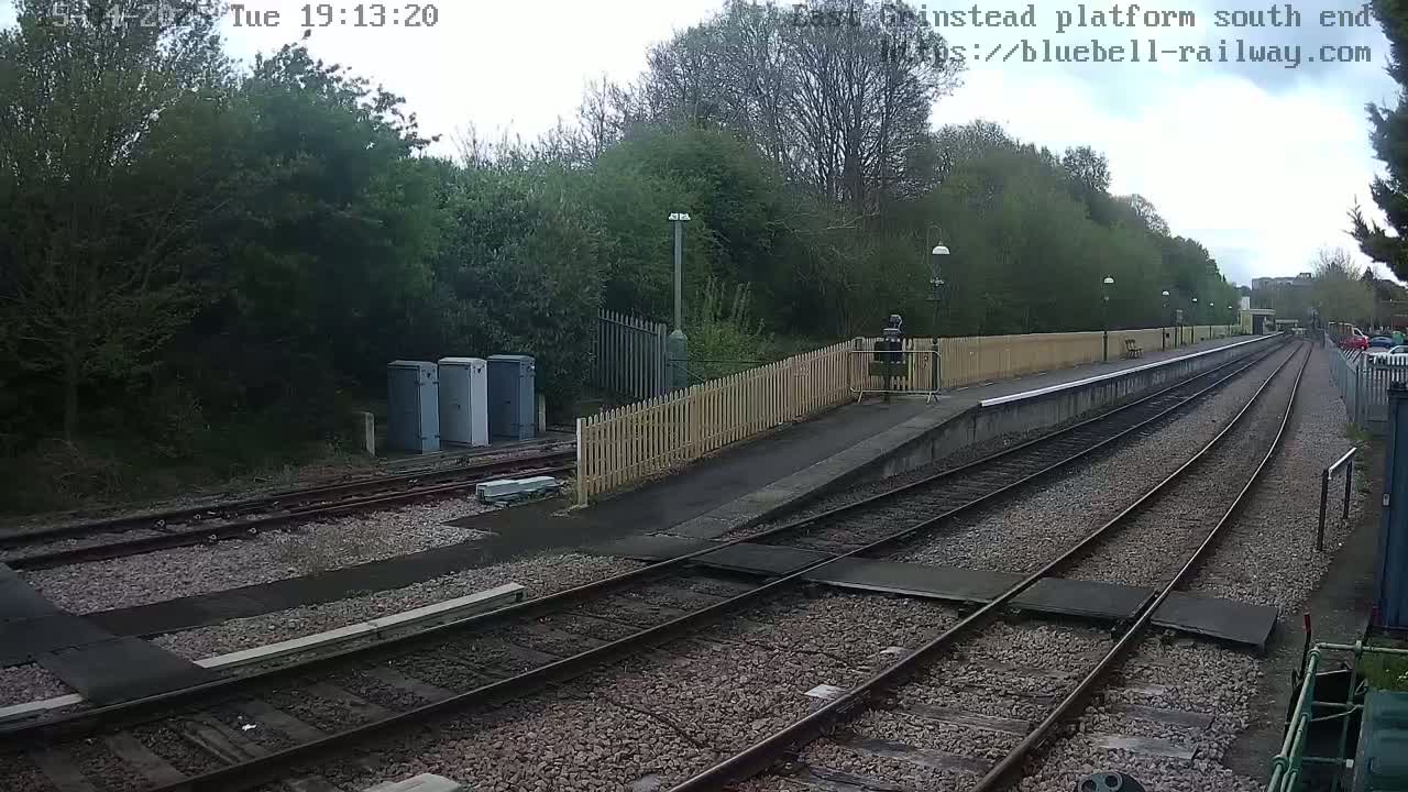 A mostly overcast view of a train platform with multiple train tracks extending into the distance, bordered by a wooden fence and lush green trees.