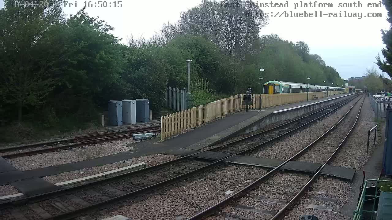 A train sits at a station platform next to a wooded area on an overcast day.